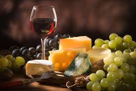 Depicted is glass of red wine in the background with different types of cheeses in the front along with grapes to highlighting this club also sells artisan cheese.